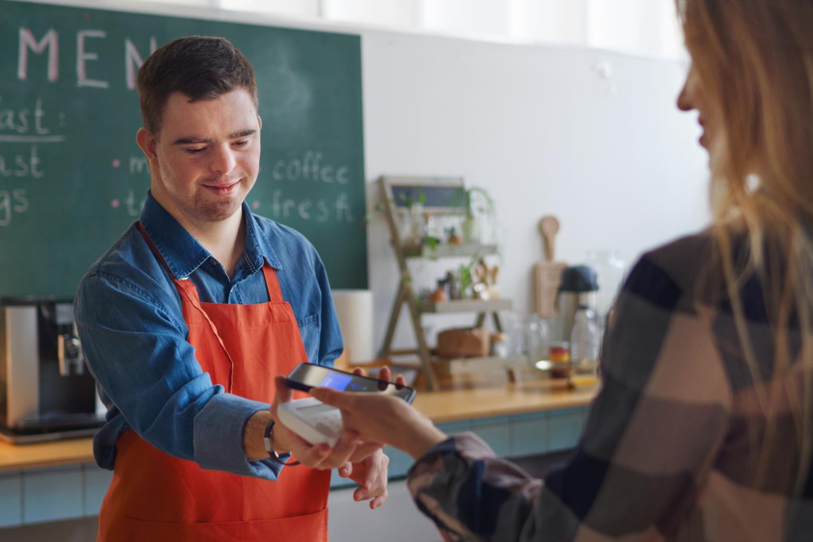 Cheerful young Down Syndrome waiter taking contactless smartphone payment from costumer in take away restaurant. stock photo