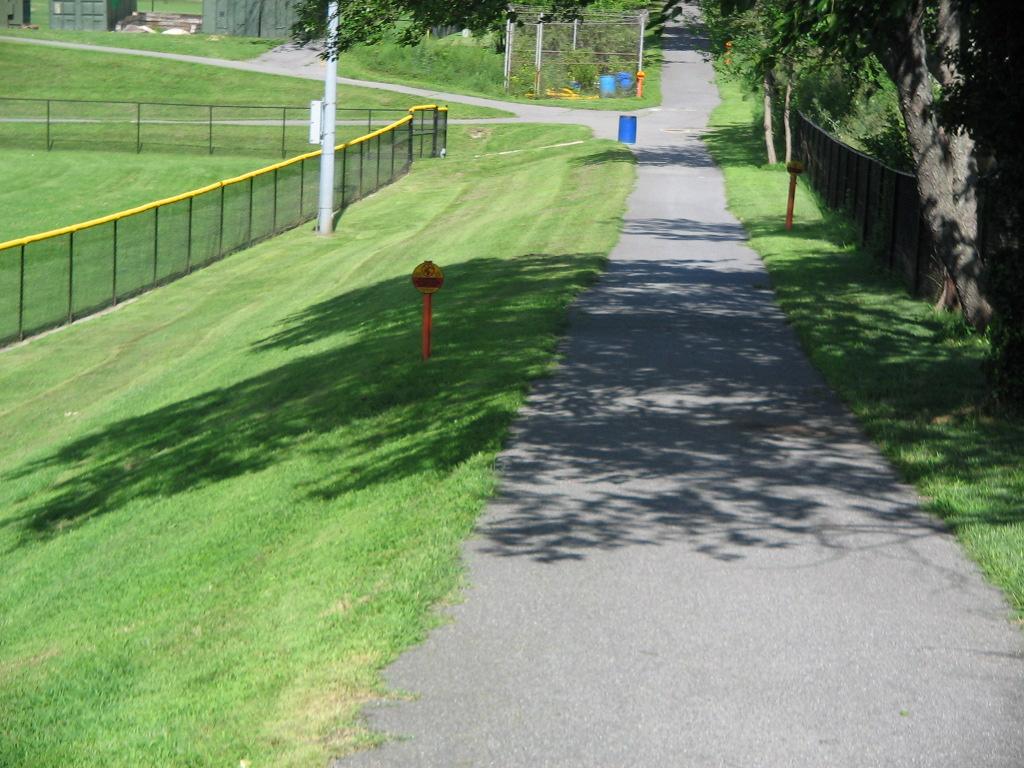Photo shows paved trail with green grass on both sides of the trail. There are shadows of trees and fence