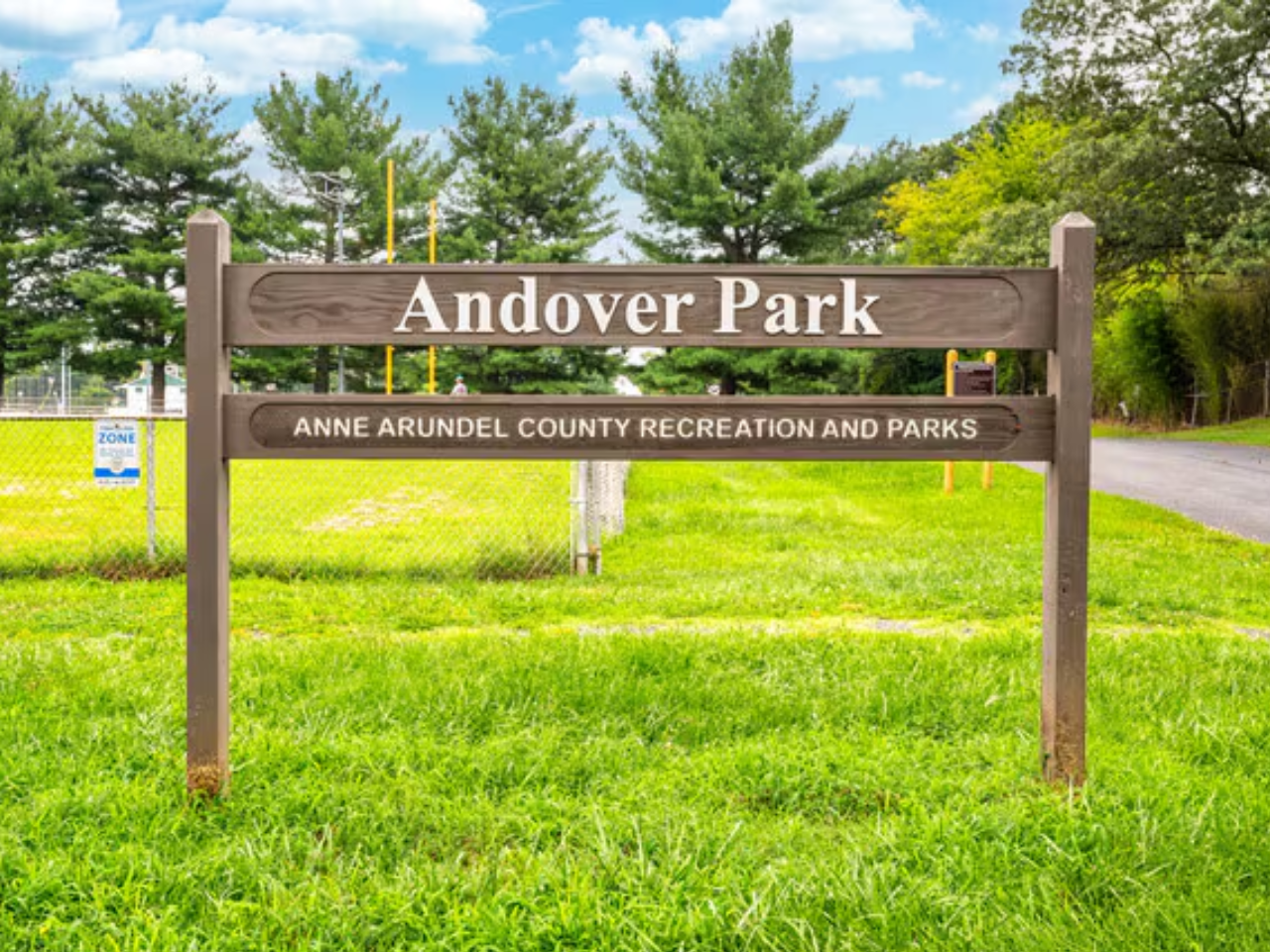 A brown, two-rail wooden sign stands on a grassy lawn under a partly cloudy sky, marking the entrance to a park. The top, wider rail features the name "Andover Park" carved in white letters. The bottom, narrower rail reads "ANNE ARUNDEL COUNTY RECREATION AND PARKS" in a smaller white font. In the background, a chain-link fence separates the lawn from an athletic field, surrounded by tall green deciduous and evergreen trees, with a paved path visible on the right.