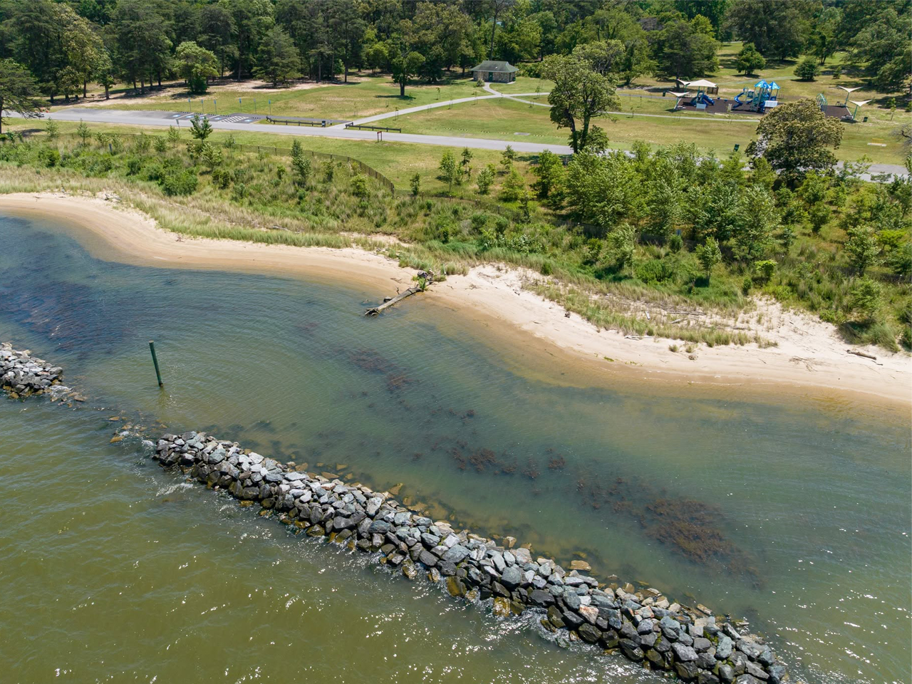 Sandy shoreline and calm water at Fort Smallwood Park under a partly cloudy sky, with a grassy bank and treeline in the background.