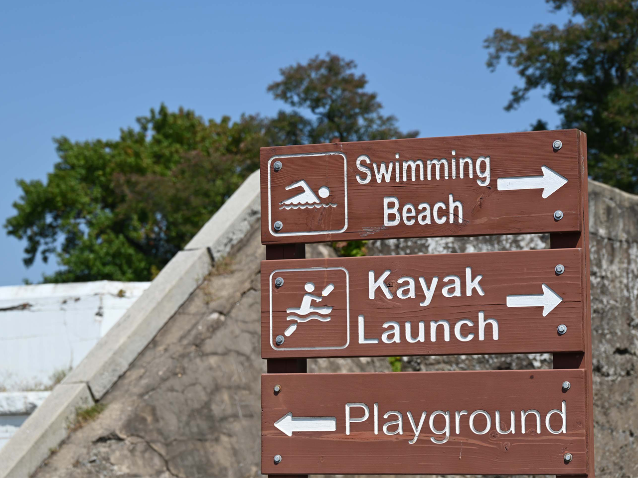 A brown, wooden directional signpost stands outdoors against a blurred background of trees and a concrete structure, featuring three distinct panels with white lettering and icons: the top panel points to the right and features an icon of a person swimming, followed by the text "Swimming Beach"; the middle panel also points to the right and displays an icon of a person in a kayak, followed by the text "Kayak Launch"; and the bottom panel points to the left and simply reads "Playground".