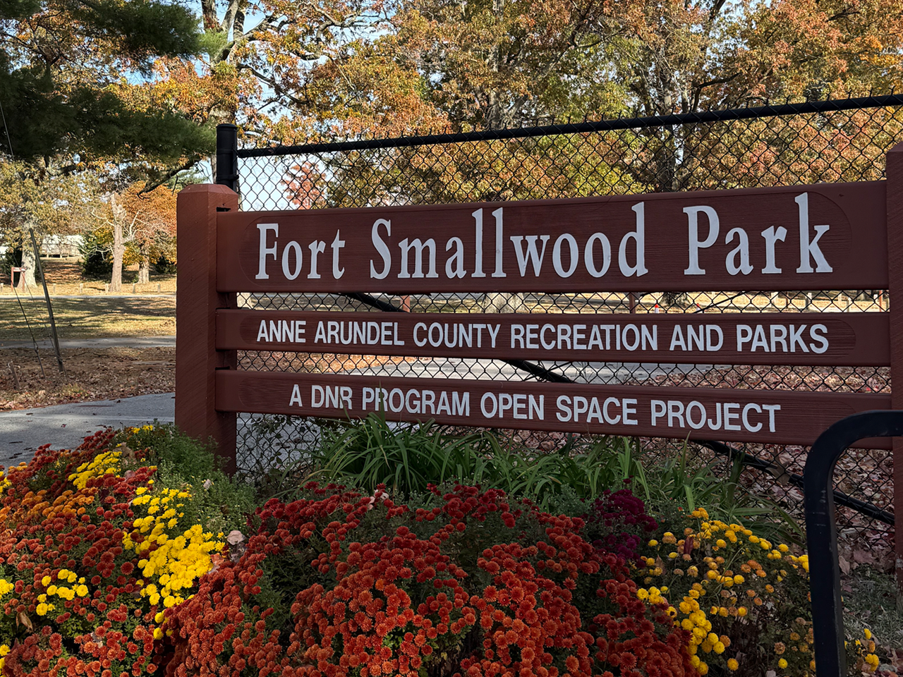 A brown wooden entrance sign reads "Fort Smallwood Park," "Anne Arundel County Recreation and Parks," and "A DNR Program Open Space Project." The sign is situated in front of a chain-link fence, with colorful fall mums in the foreground.