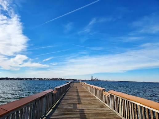 A wooden fishing pier stretches into calm, blue water under a bright sky with wispy clouds and contrails. The pier features railings on both sides, leading the eye toward the distant shoreline visible on the horizon under the expansive blue sky.