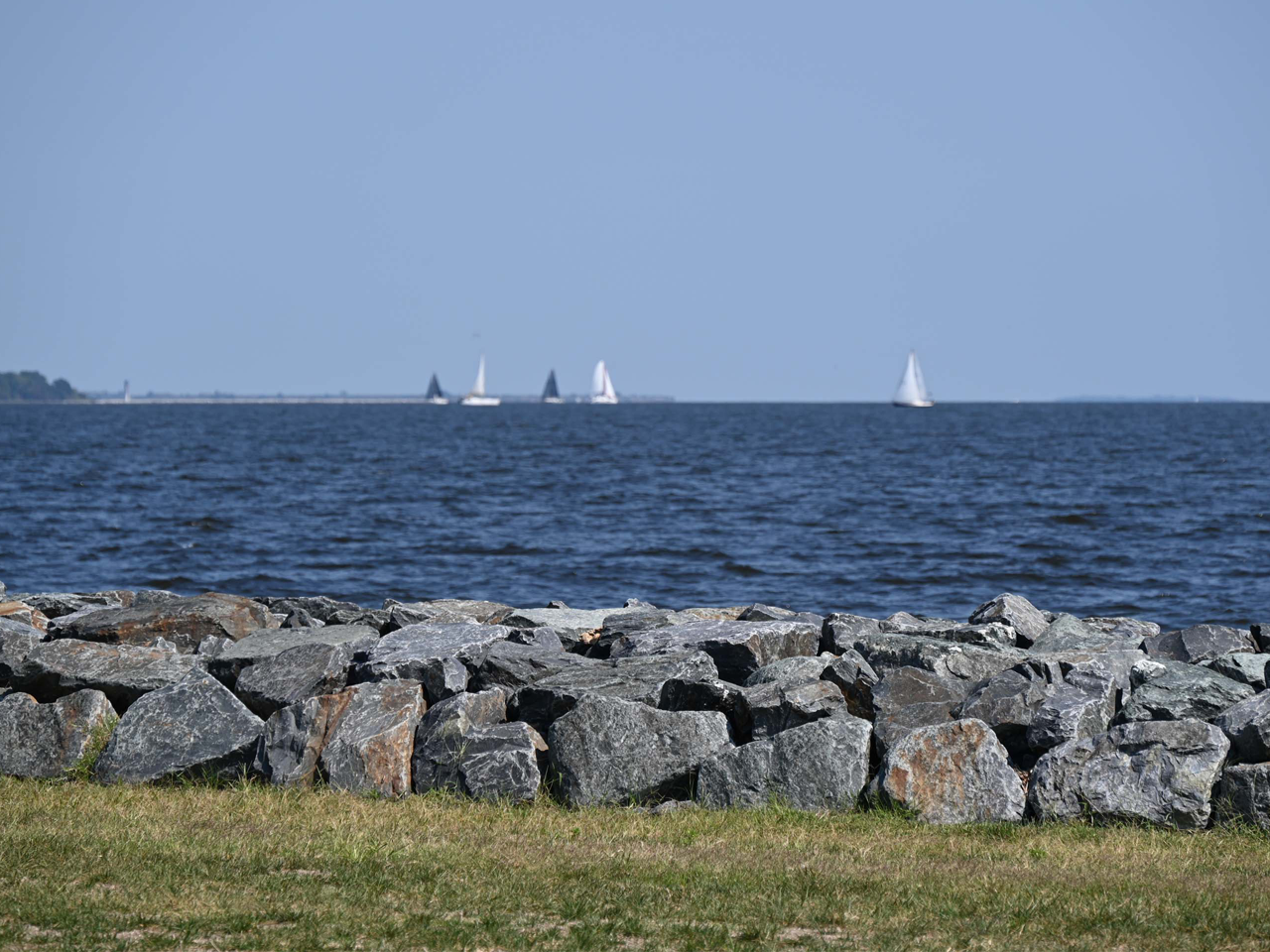 A scenic, wide-angle shot taken from the shoreline of Fort Smallwood Park captures a tranquil view across the deep blue waters of the Patapsco River on a clear, sunny day. In the immediate foreground, a jagged stone bulkhead made of large, grey, weathered rocks stretches horizontally across the bottom of the frame, separating a thin strip of dry, golden-green grass from the water's edge. Beyond the stone barrier, the expansive river dominates the middle and background.