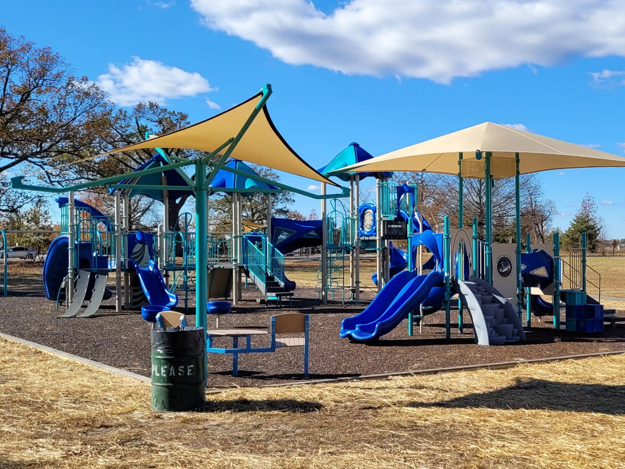 Outdoor playground with blue and teal play structures, covered by large tan shade sails, set on a mulch bed under a partly cloudy blue sky. A green trash can with "PLEASE" written on it sits in the foreground.
