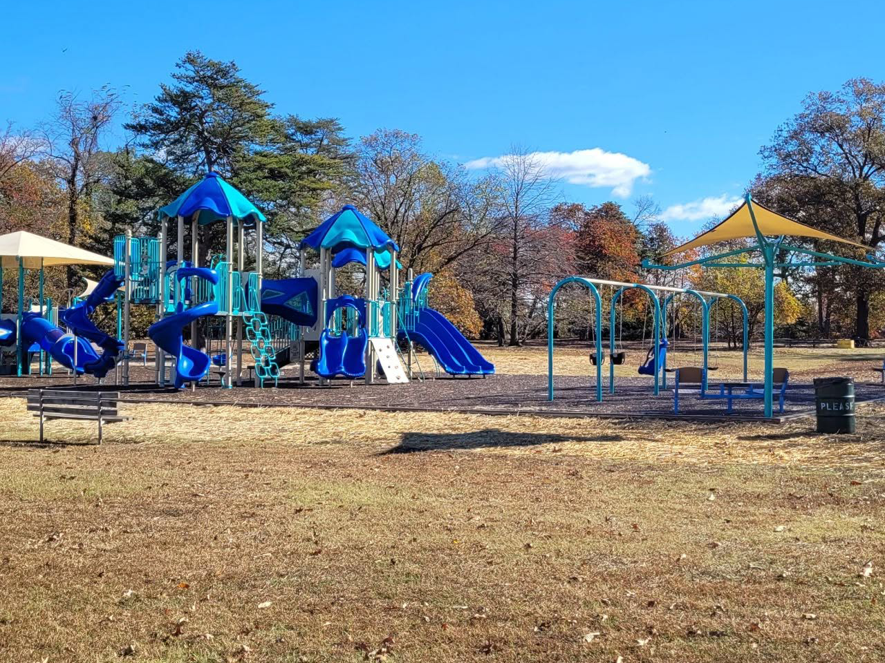 Outdoor playground with blue and teal play structures, covered by large tan shade sails, set on a mulch bed under a partly cloudy blue sky. A green trash can with "PLEASE" written on it sits in the foreground.