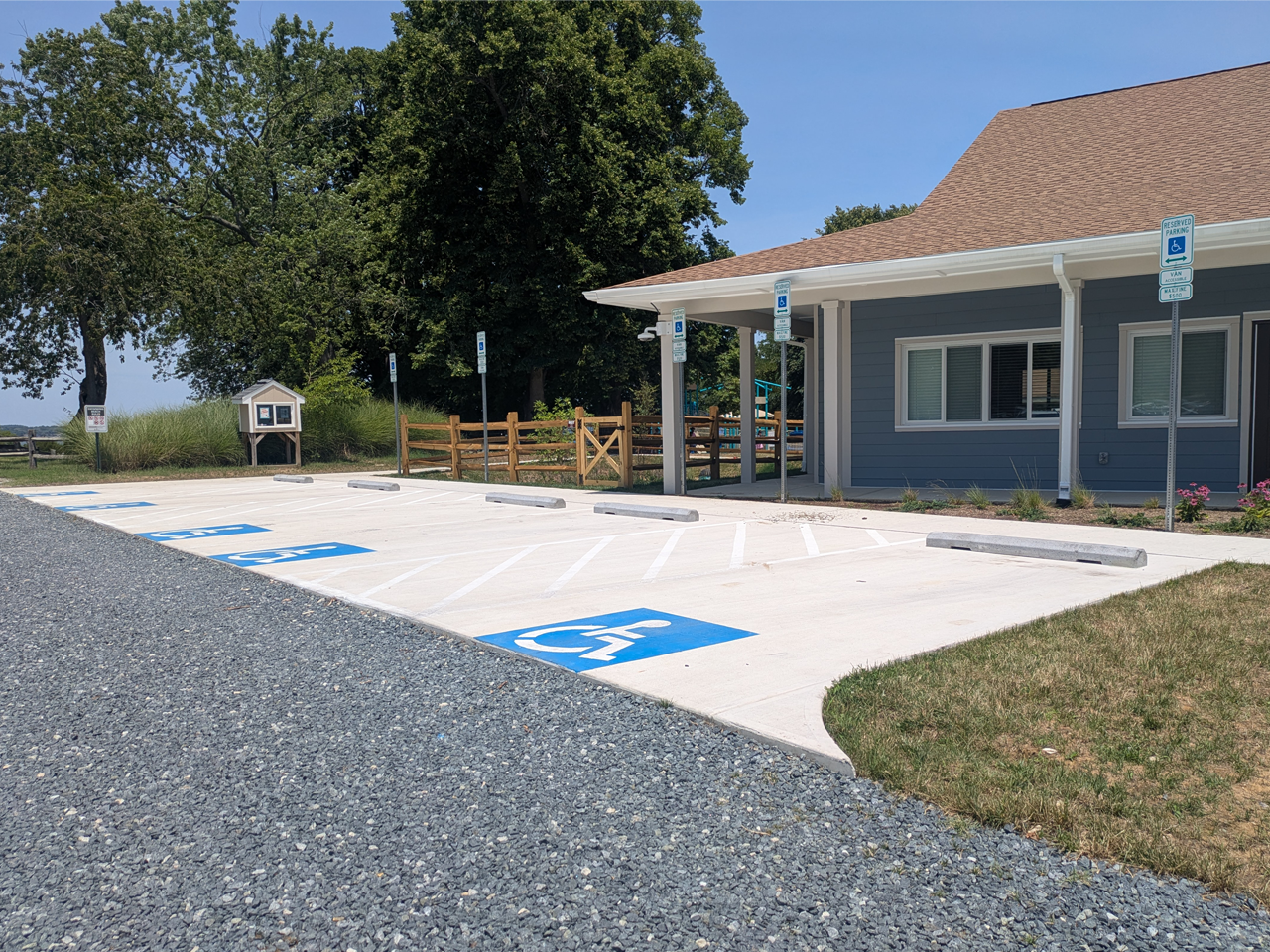 A newly paved concrete accessible parking area sits next to a single-story blue building with a brown shingled roof. The lot features designated spaces painted with blue and white wheelchair icons and white diagonal crosshatch loading zones. Each space has a concrete wheel stop and a metal signpost, including a "Van Accessible" sign. In the foreground, a gray gravel driveway leads to the spaces. In the background, a wooden split-rail fence separates the lot from a grassy park with trees and a kiosk.