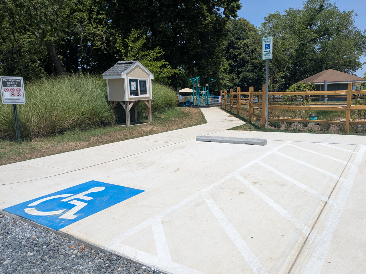 A newly paved concrete accessible parking space and an adjacent white-striped loading zone. A concrete wheel stop sits at the head of the parking space, which is marked by a green "Reserved Parking" sign with a wheelchair icon. Just past the parking space, a paved concrete walkway leads toward a park area. To the left of the walkway is a small, elevated wooden kiosk and a "Swimming Beach Rules" sign. To the right, a wooden split-rail fence runs along a landscaped area, with a playground structure.