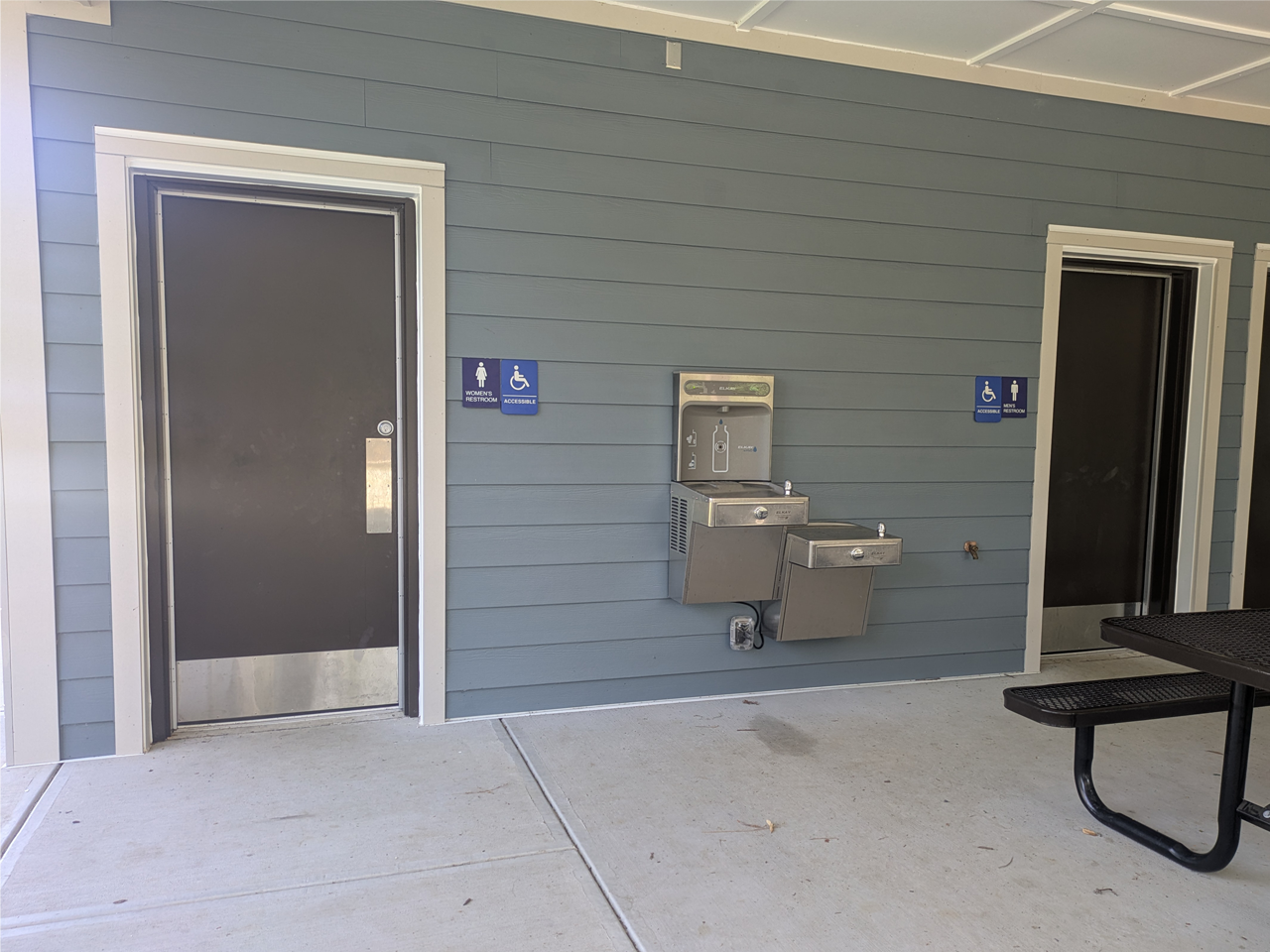 An outdoor, covered pavilion features a blue-gray horizontal siding wall with two dark metal doors and a stainless-steel water fountain station. The door has a blue "Women's Restroom" sign and a blue "Accessible" wheelchair sign. To the right, doorway leads into the men's restroom, marked by a matching set of blue signs. Between the two restroom entrances is a dual-level water fountain unit, complete with a water bottle filling station. The concrete ground extends under the pavilion.