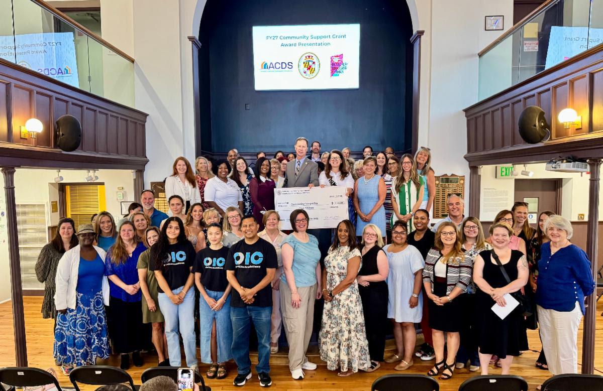 A large group of diverse people stands together indoors in a room with wood-paneled walls, posing for a photo during an FY27 Community Support Grant Award Presentation. A screen in the background displays the event title and organization logos. Several people in the front row are holding a large ceremonial check for $100,000.