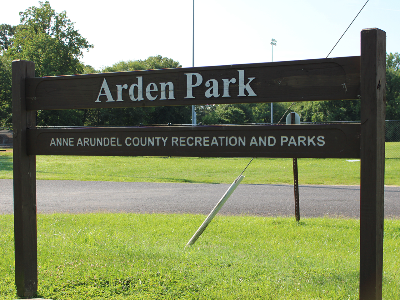 A dark brown wooden sign mounted on two posts sits in the grass, displaying white lettering that reads "Arden Park" on the top rail and "ANNE ARUNDEL COUNTY RECREATION AND PARKS" on the bottom rail. In the background, a paved road stretches past the sign toward a bright green lawn bordered by tall trees under a sunny sky.