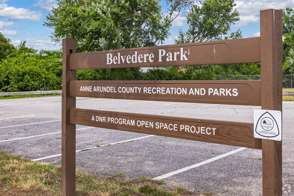 A brown wooden entrance sign with white lettering stands in the foreground, reading "Belvedere Park," followed by "ANNE ARUNDEL COUNTY RECREATION AND PARKS," and "A DNR PROGRAM OPEN SPACE PROJECT." A small white "Land & Water Conservation Fund" logo emblem is attached to the bottom right post. Behind the sign, an empty, paved parking lot with white painted stalls stretches toward lush green trees under a bright, partly cloudy sky.