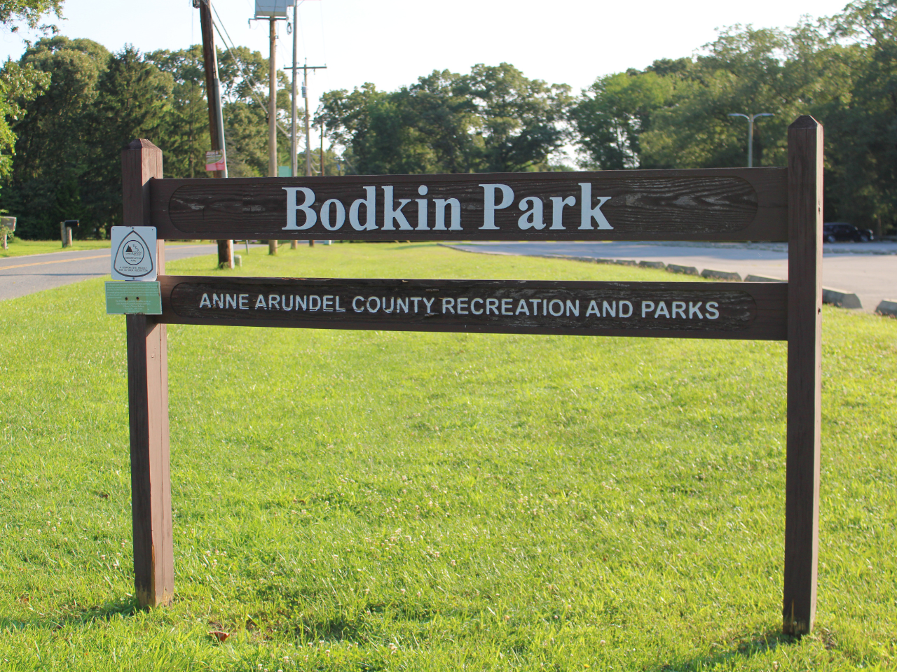 A dark brown wooden sign with white carved text stands on a grassy lawn, reading "Bodkin Park" on the top rail and "ANNE ARUNDEL COUNTY RECREATION AND PARKS" on the bottom rail. A small white Land & Water Conservation Fund emblem is attached to the left post. In the background, a paved road sits to the left, while an open parking lot and dense green trees stretch behind the sign under bright daylight.