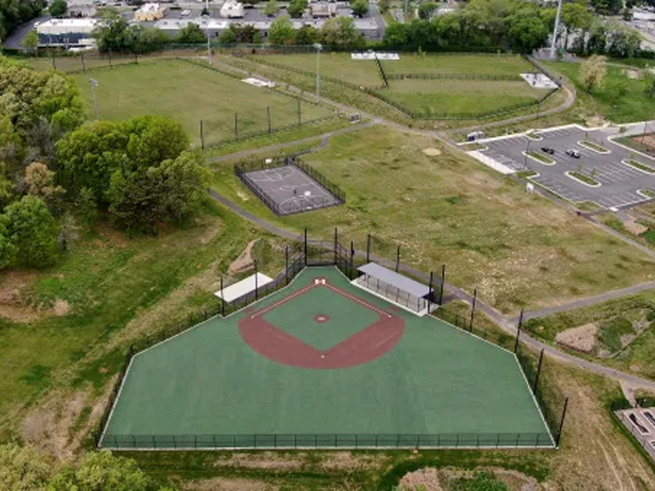 An aerial photograph shows a newly constructed adaptive baseball/softball field featuring a smooth green surface, reddish-brown baseline paths, covered dugouts, and black chain-link perimeter fencing. Surrounding the field are paved walking paths, an outdoor basketball court, a paved parking lot, fenced community garden plots, and adjacent grassy park fields bordered by lush green trees.