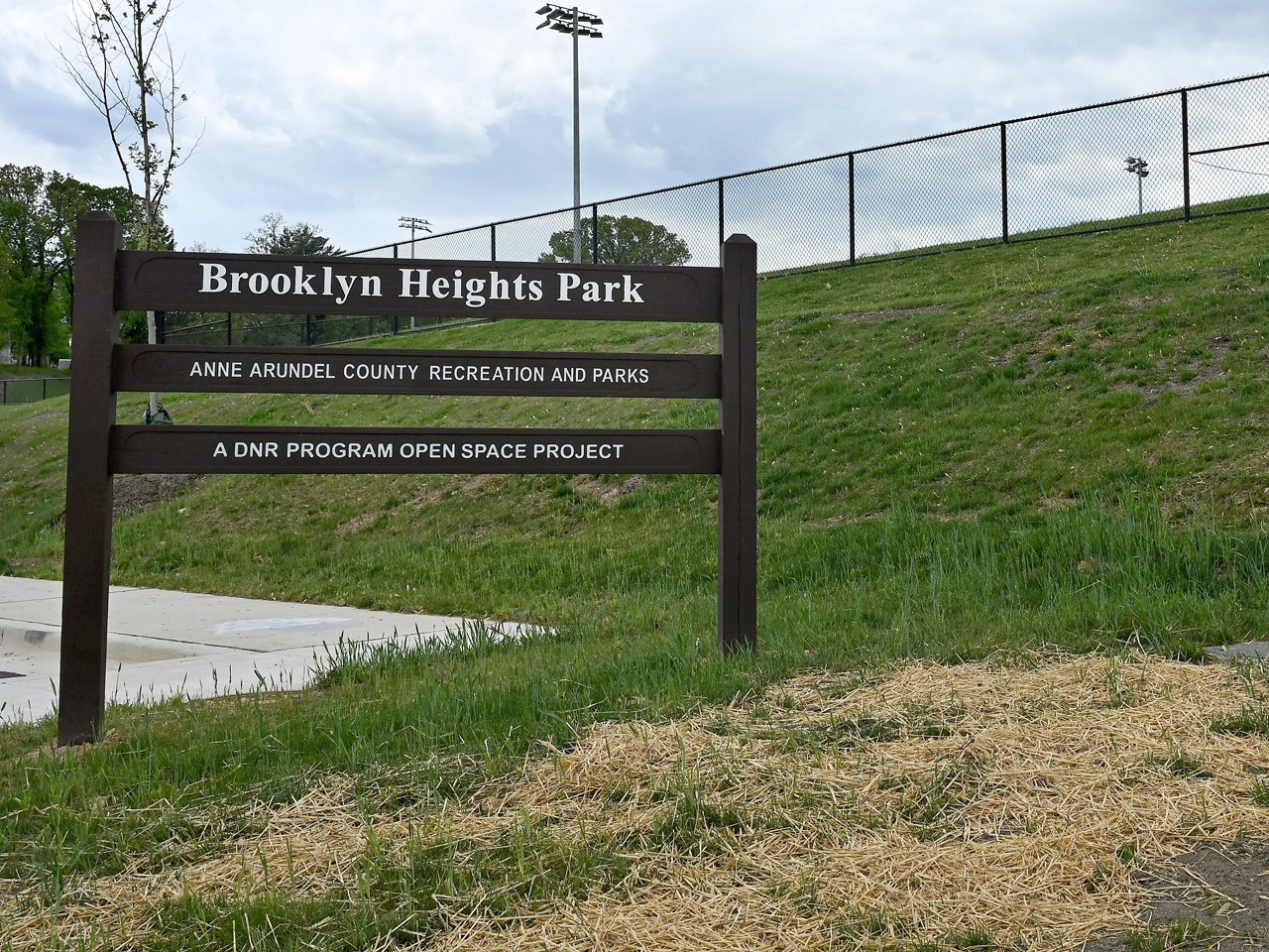 A brown wooden sign stands in the foreground on a grassy patch covered with scattered straw, displaying white text that reads "Brooklyn Heights Park," followed by "ANNE ARUNDEL COUNTY RECREATION AND PARKS," and "A DNR PROGRAM OPEN SPACE PROJECT." Behind the sign, a grassy slope rises toward a black chain-link fence running along the crest of the hill under an overcast, cloudy sky.