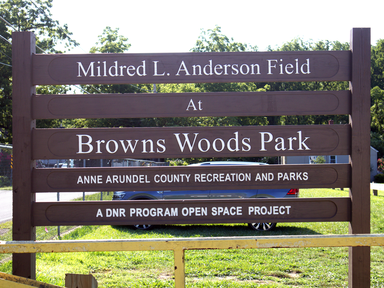 A dark brown wooden sign mounted on two posts displays white carved lettering across five horizontal rails: "Mildred L. Anderson Field," "At," "Browns Woods Park," "ANNE ARUNDEL COUNTY RECREATION AND PARKS," and "A DNR PROGRAM OPEN SPACE PROJECT." In the background, a blue vehicle is parked on a grassy patch near a chain-link fence and green trees under bright daylight, with a yellow metal barrier visible in the immediate foreground.