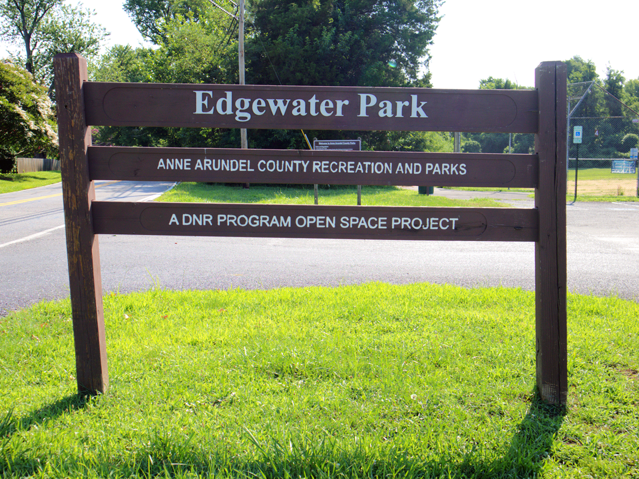 A dark wooden sign standing in a grassy area that reads "Edgewater Park" at the top, followed by "ANNE ARUNDEL COUNTY RECREATION AND PARKS" and "A DNR PROGRAM OPEN SPACE PROJECT" on three horizontal slats. 