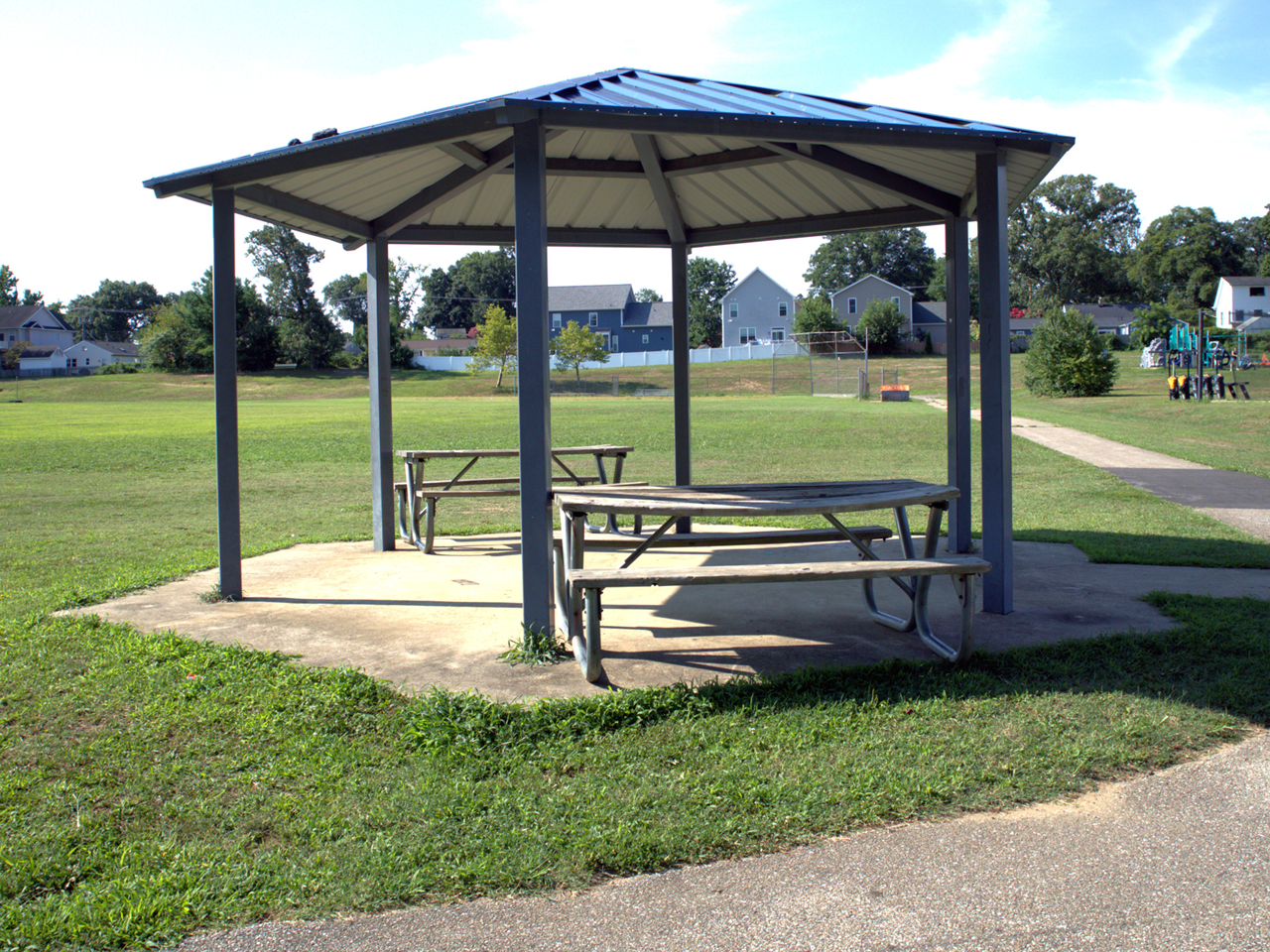 Outdoor gazebo on pavement with two picnic tables, green grass and bright blue skies. 