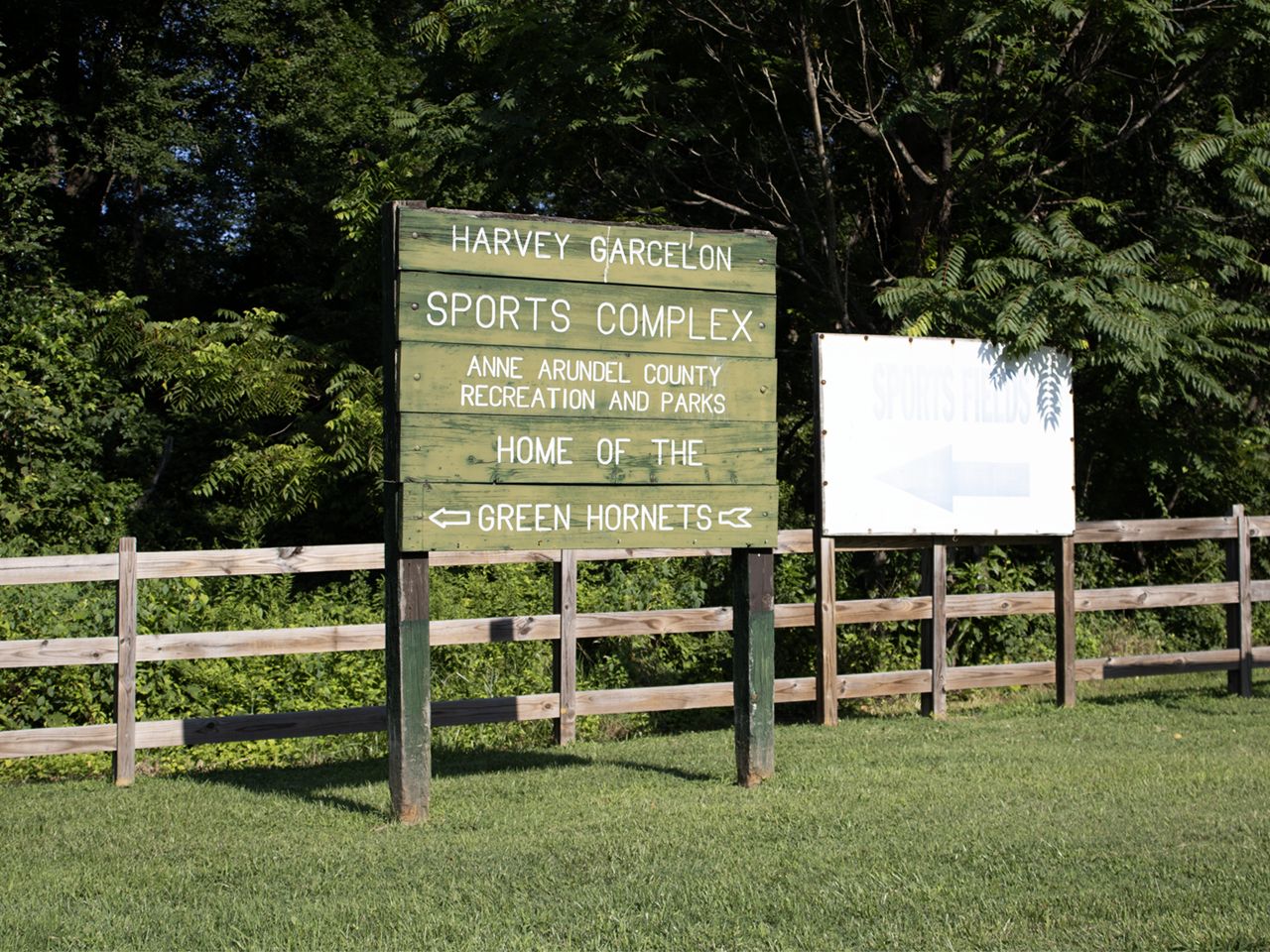 A green painted wooden entrance sign with white lettering stands on a lush lawn, reading "HARVEY GARCELON SPORTS COMPLEX," "ANNE ARUNDEL COUNTY RECREATION AND PARKS," and "HOME OF THE GREEN HORNETS" with double-sided arrows. Behind it, a wooden three-rail fence runs past a white sign pointing left toward "SPORTS FIELDS," set against a backdrop of dense green trees and foliage under bright daylight.