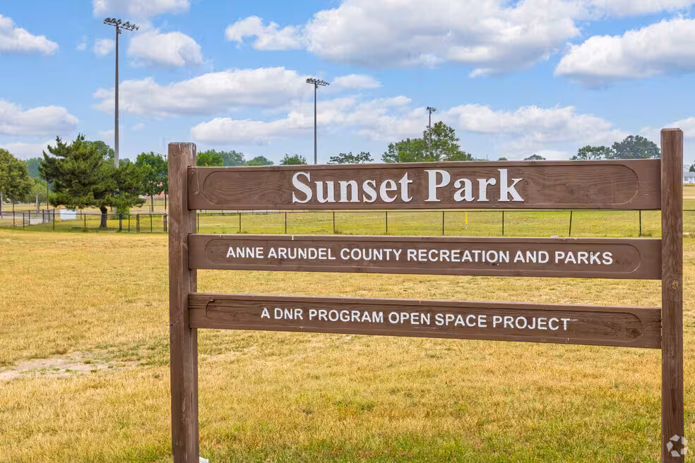 A wooden post-and-beam sign standing in a dry, grassy field under a partly cloudy blue sky. The top panel reads "Sunset Park" in large white lettering, followed by "Anne Arundel County Recreation and Parks" and "A DNR Program Open Space Project." In the background, chain-link fencing, open athletic fields, and tall floodlight poles are visible. 
