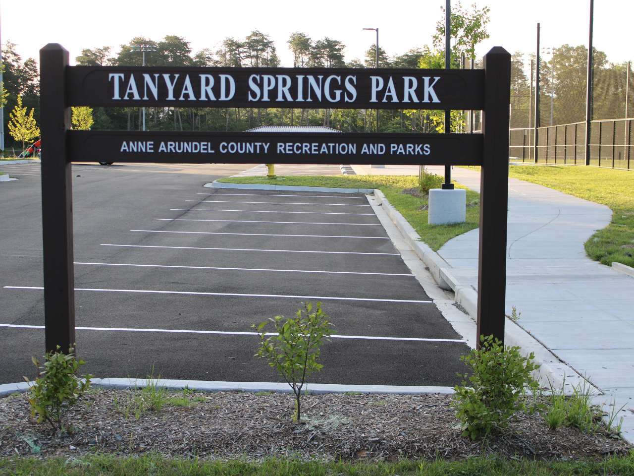 A dark wooden entrance sign for "TANYARD SPRINGS PARK" stands prominently in the foreground, with smaller white text underneath reading "ANNE ARUNDEL COUNTY RECREATION AND PARKS". Small green bushes planted in mulch sit right in front of the sign. Behind the sign, an empty, freshly paved asphalt parking lot with bright white painted parking spaces stretches into the background. To the right of the parking lot, a concrete sidewalk curves past a grassy strip toward sports fields enclosed by tall fencing, with