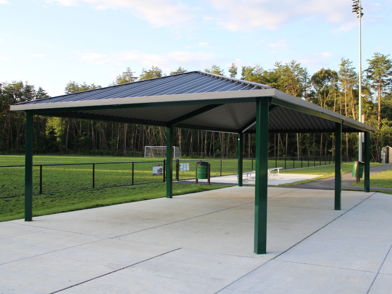 A large covered park pavilion with a dark metallic gabled roof supported by green steel posts sits on a concrete pad. Underneath and adjacent to the shelter, a green trash receptacle and a white bleacher bench are visible. In the background beyond a black chain-link fence, an athletic field with a soccer goal is framed by a dense line of trees under a light sky with a tall stadium light pole standing nearby.