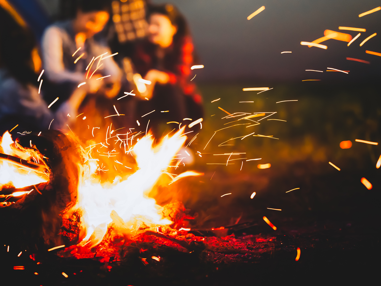 Sparking bonfire with people sit around bright bonfire near camping tent in forest in summer night background. Group of student at outdoor fire fuel. 