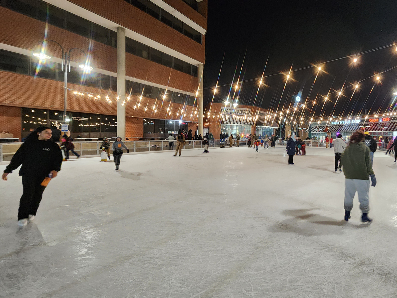 People ice skating at an outdoor rink illuminated by warm string lights at night, surrounded by brick town center buildings including a pharmacy and clock tower.
