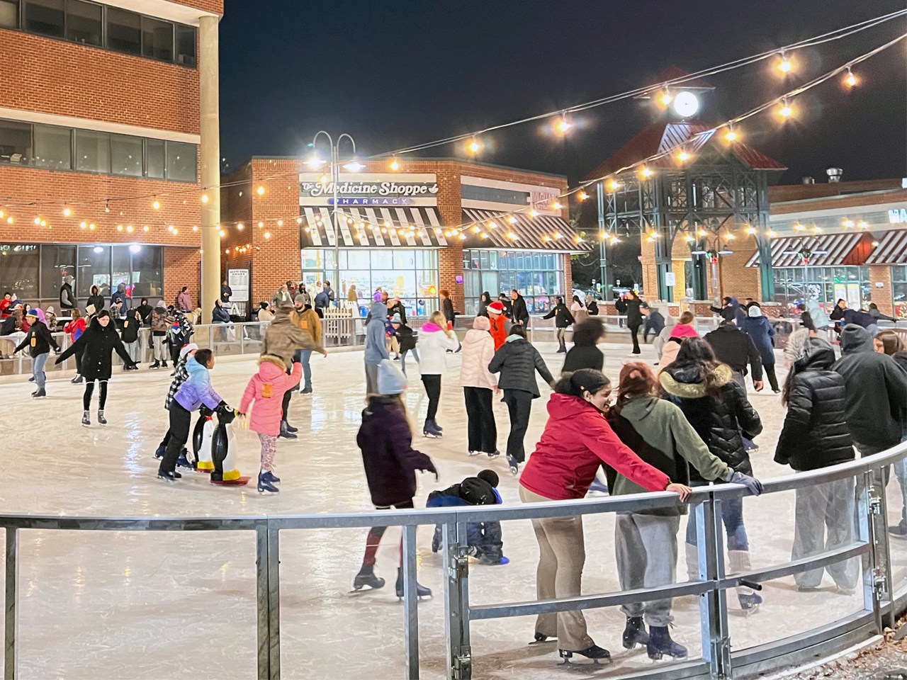 People ice skating on a wide outdoor rink at night under glowing string lights, framed by brick town center buildings and local storefronts.