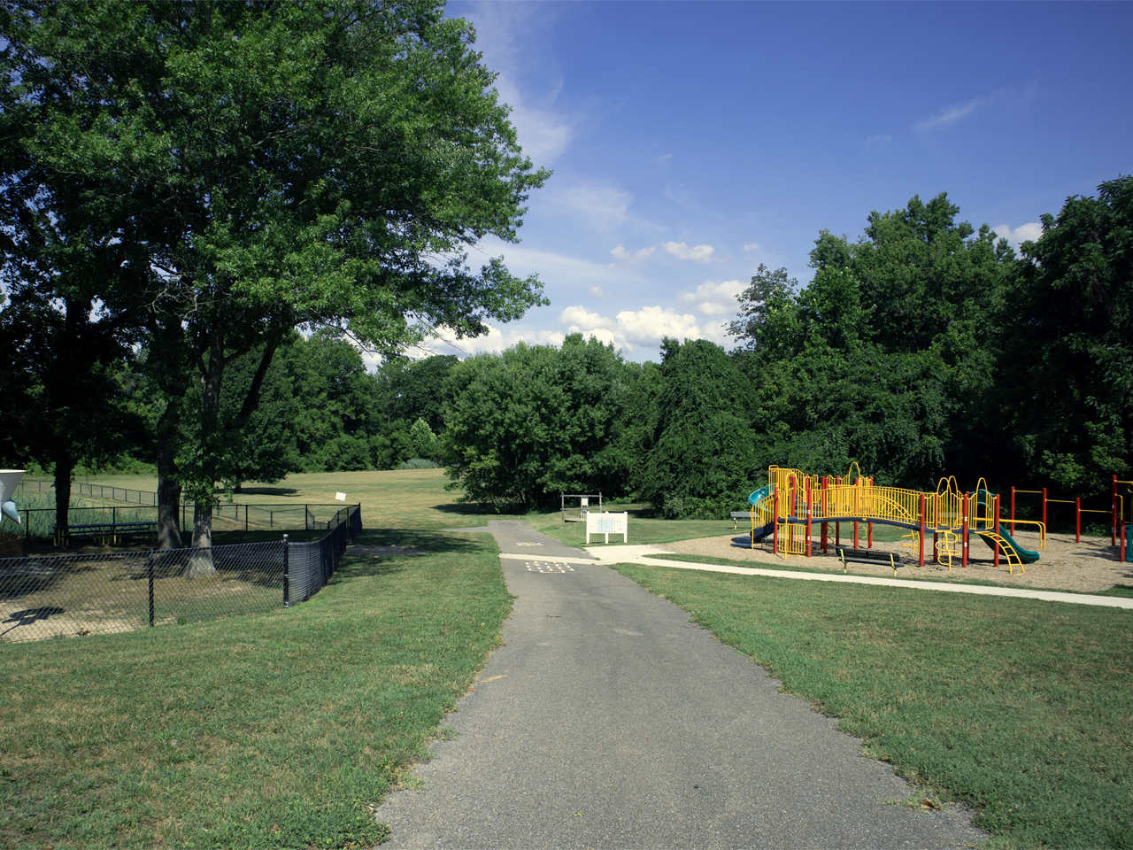 A paved asphalt walking path runs down the center of a grassy park scene towards open fields and dense green trees. On the right, a colorful children's playground structure with red, yellow, and green slides and climbing equipment sits in a woodchip bed. To the left, a chain-link fence encloses a shaded area beneath large, leafy mature trees under a clear blue sky with light clouds.