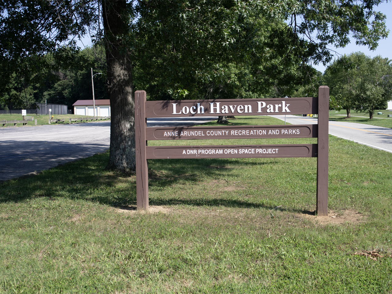 A brown wooden entrance sign mounted on two posts stands on a grassy lawn beneath the shade of a large tree, featuring white text across three horizontal rails reading "Loch Haven Park," "ANNE ARUNDEL COUNTY RECREATION AND PARKS," and "A DNR PROGRAM OPEN SPACE PROJECT." To the left of the sign, an asphalt parking lot stretches toward park buildings in the background, while a two-lane paved road runs along the right side next to residential homes under a clear blue sky.