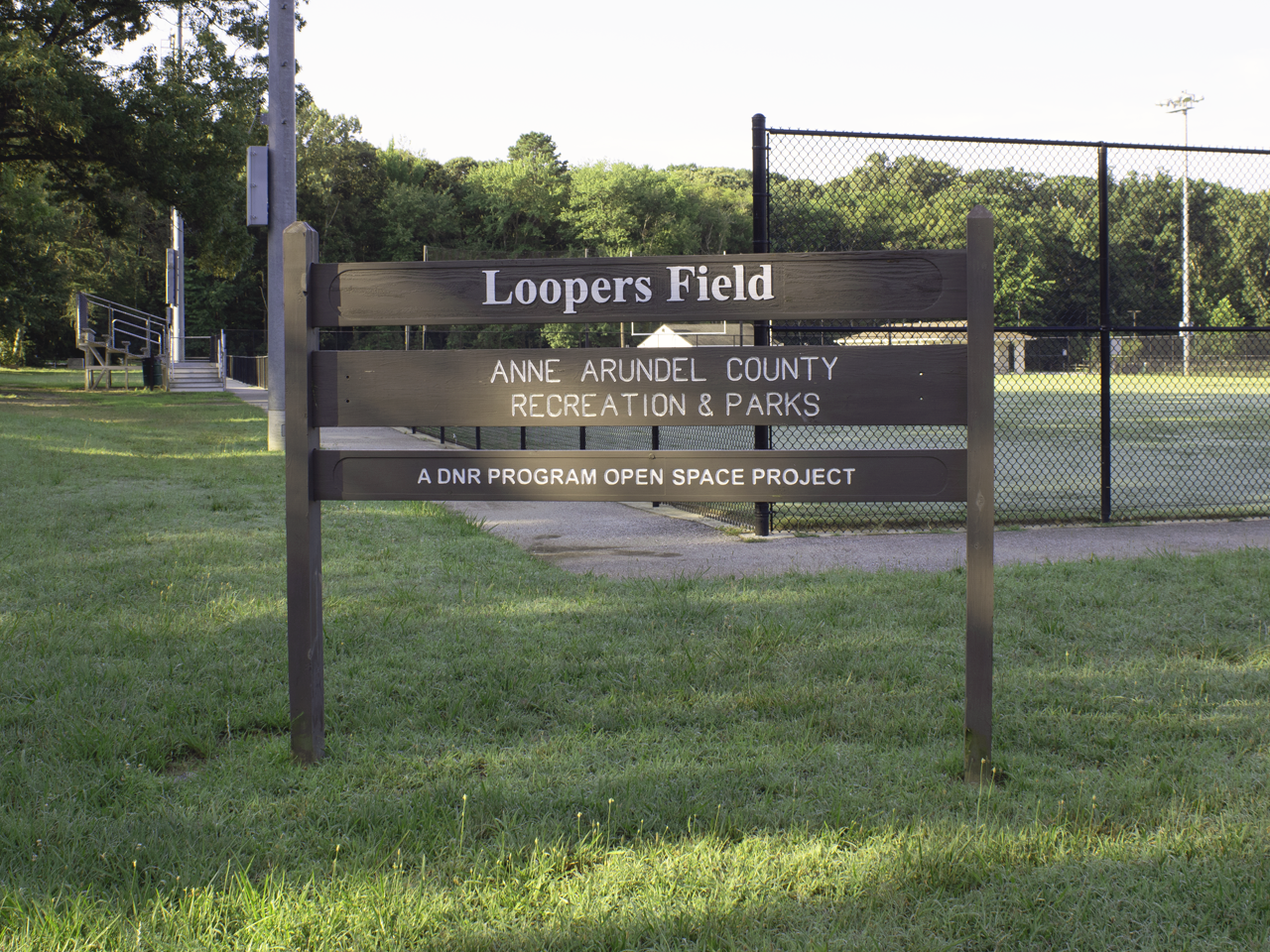 A wooden park sign reading "Loopers Field, Anne Arundel County Recreation & Parks, A DNR Program Open Space Project" standing in front of a fenced athletic field.