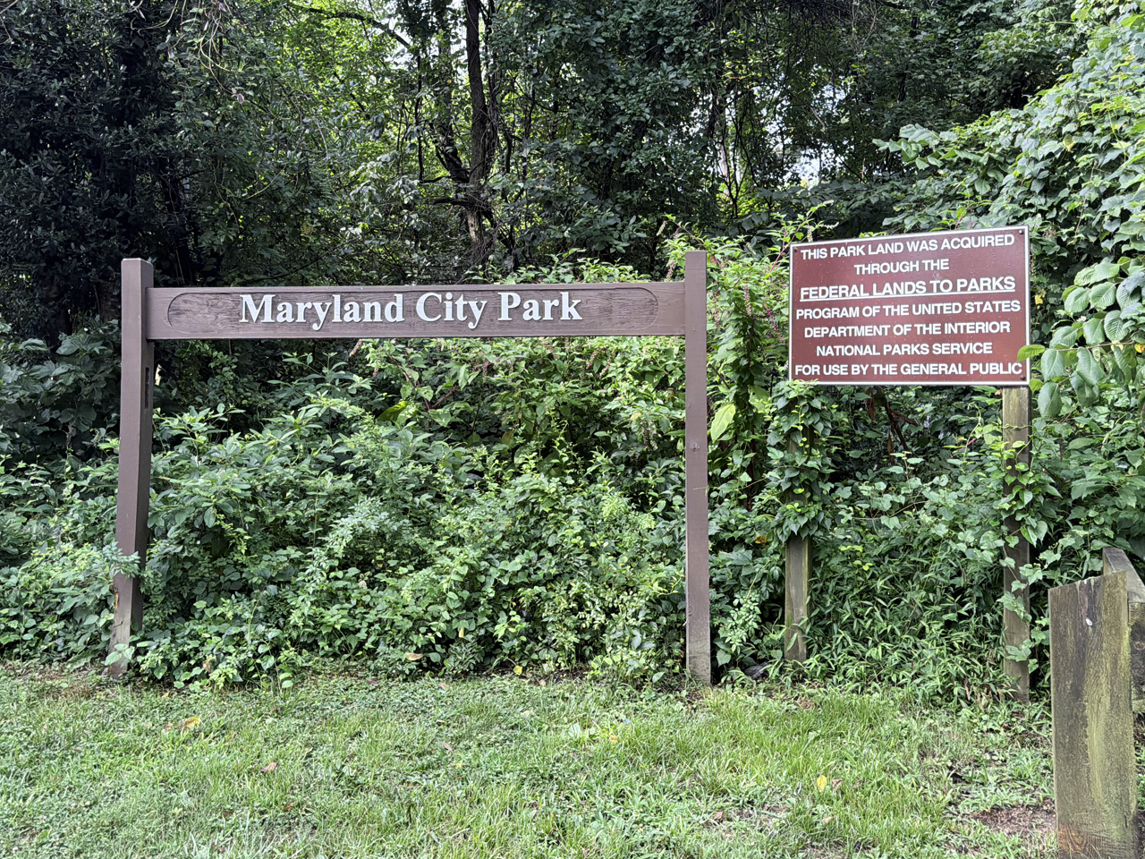 A wooden post-and-beam park sign reading "Maryland City Park" against a lush, overgrown backdrop of green trees and bushes. To the right, a second brown sign reads: "This park land was acquired through the Federal Lands to Parks Program of the United States Department of the Interior National Parks Service for use by the general public.