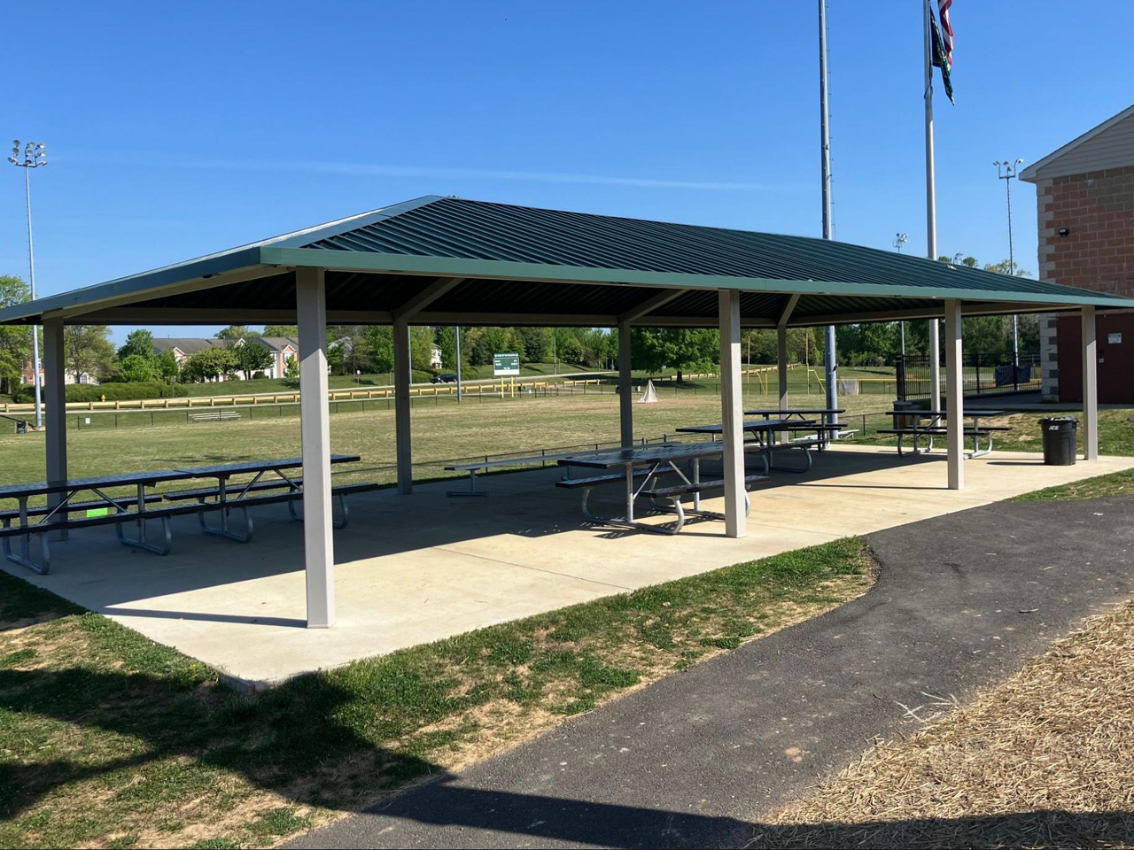 A spacious, open-air park pavilion features a dark green metal roof supported by off-white square posts set upon a concrete slab. Underneath the structure, several grey metal picnic tables with attached benches sit in the shade. An asphalt pathway curves along the right side of the pavilion next to a patch of straw, leading past a black trash receptacle toward a brick park facility building. In the background, tall flagpoles fly the American and state flags against a clear and bright blue sky.