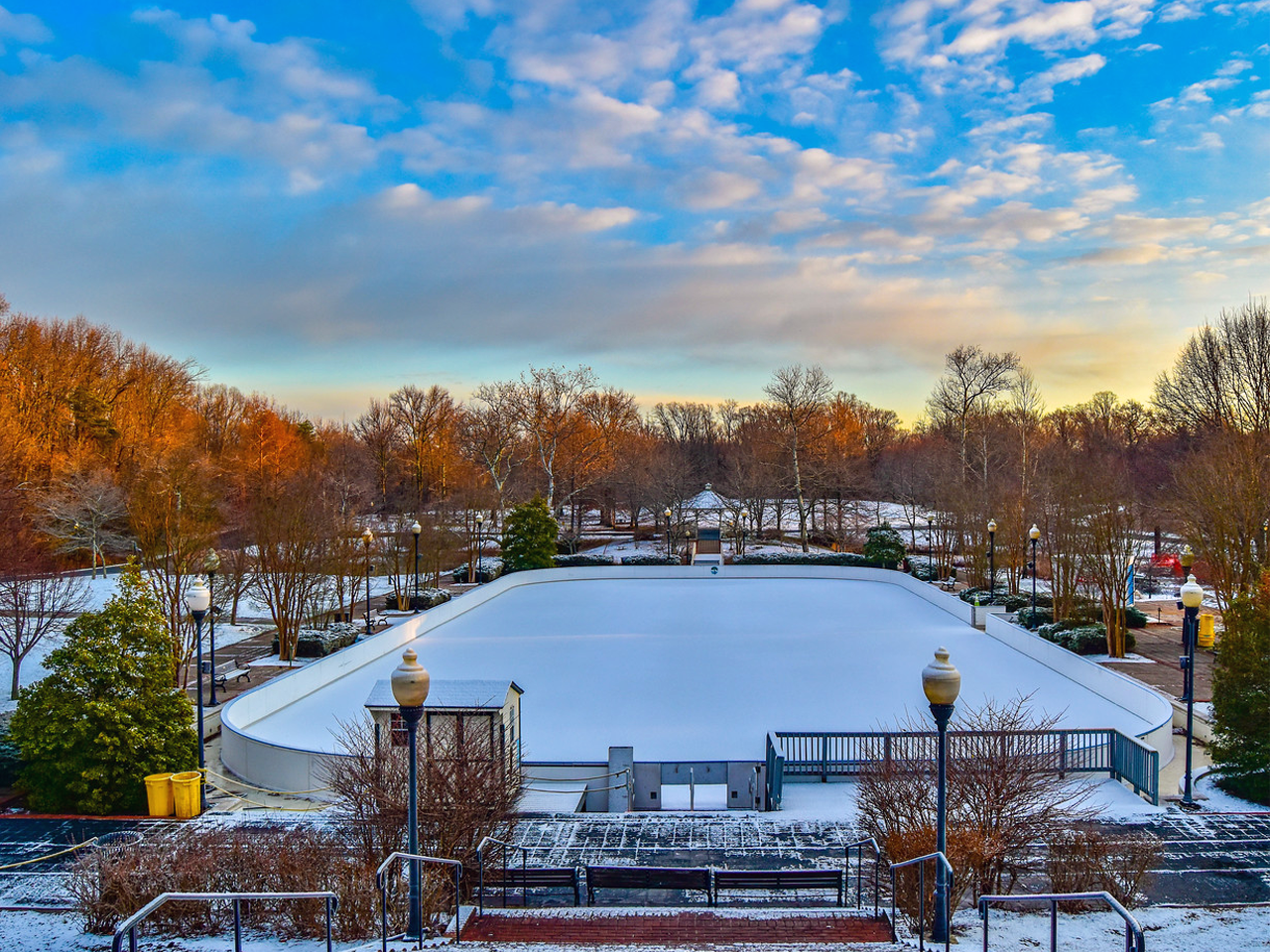 An empty, snow-dusted outdoor ice skating rink surrounded by bare winter trees, vintage lamp posts, and a bright blue sky with light clouds.