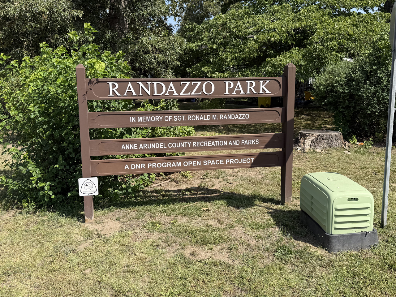 A brown wooden sign standing on a grassy area in front of green trees and foliage. The main header reads "Randazzo Park," followed by three smaller sub-lines: "In Memory of Sgt. Ronald M. Randazzo," "Anne Arundel County Recreation and Parks," and "A DNR Program Open Space Project." To the right of the sign sits a small green utility box next to a metal signpost. 