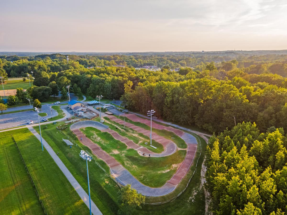 An elevated drone photo showing a dirt BMX racing track with paved turns, jumps, and berms. The track is equipped with tall floodlights, spectator bleachers, and a start hill structure. Located next to a green athletic field and parking area, the entire facility is bordered by a dense forest under a warm evening sky. 