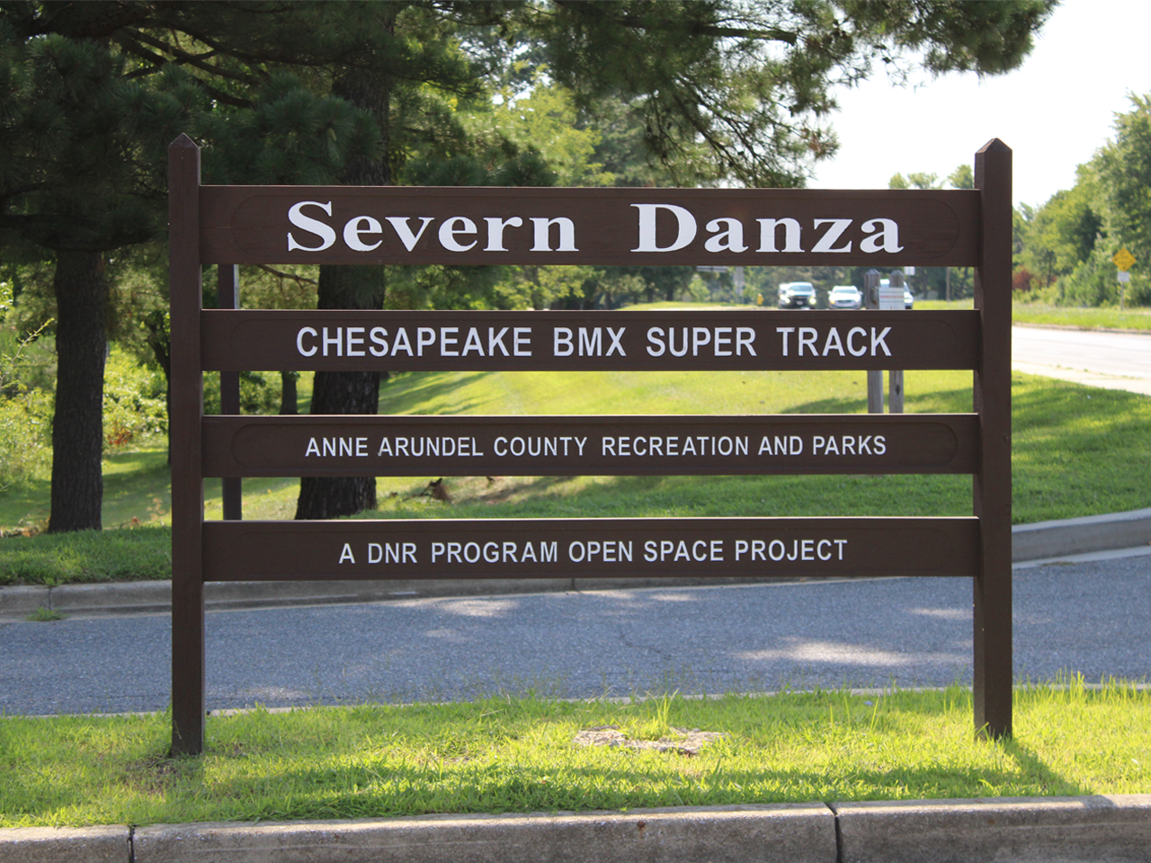 A brown wooden park sign reading "Severn Danza" and "Chesapeake BMX Super Track" set in front of a grassy lawn and trees.