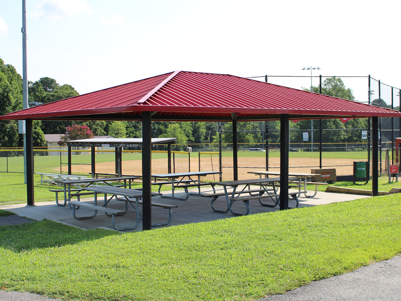 An outdoor covered pavilion featuring a red corrugated metal hip roof supported by black steel posts. Underneath the concrete pad sit multiple wooden picnic tables. In the background, a fenced baseball dirt infield, dugouts, and tall field lighting are visible against a backdrop of green trees. 
