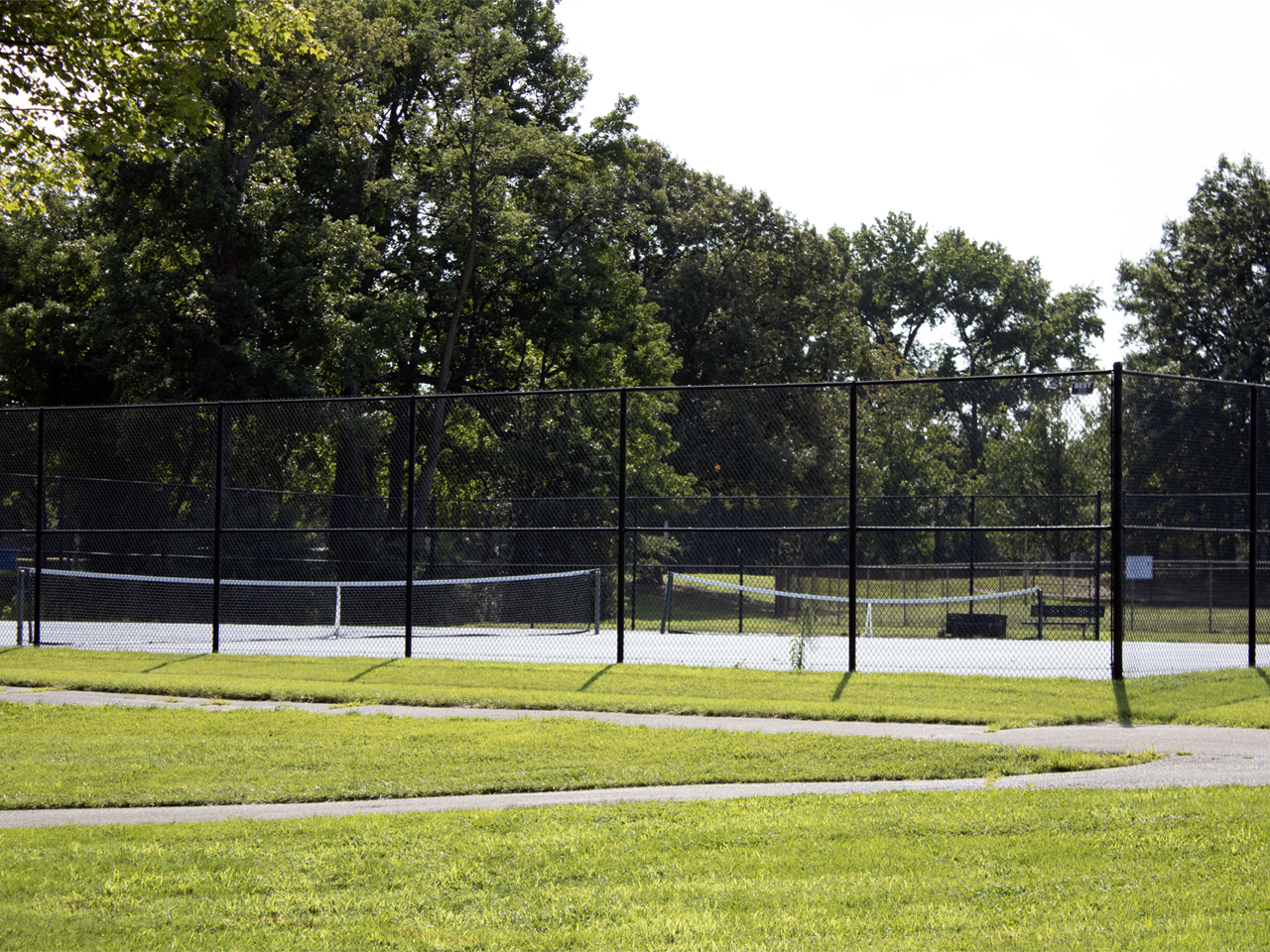 Two outdoor paved courts with white nets, enclosed by a tall black chain-link fence. In the foreground, a paved walking path curves through a bright green grassy lawn. Behind the courts, a dense line of tall, leafy green trees stands under a bright sky. A small bench is visible inside the fenced area on the right court. 