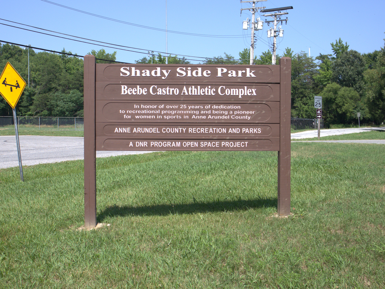 A brown post-and-beam wooden sign standing on a grassy area under a clear blue sky. The top header reads "Shady Side Park" with "Beebe Castro Athletic Complex" below it. A dedication plaque in the middle honors over 25 years of dedication to recreational programming for women in sports in Anne Arundel County, followed by text for Anne Arundel County Recreation and Parks and the DNR Program Open Space Project. To the left stands a yellow diamond-shaped sign depicting children on a seesaw. 