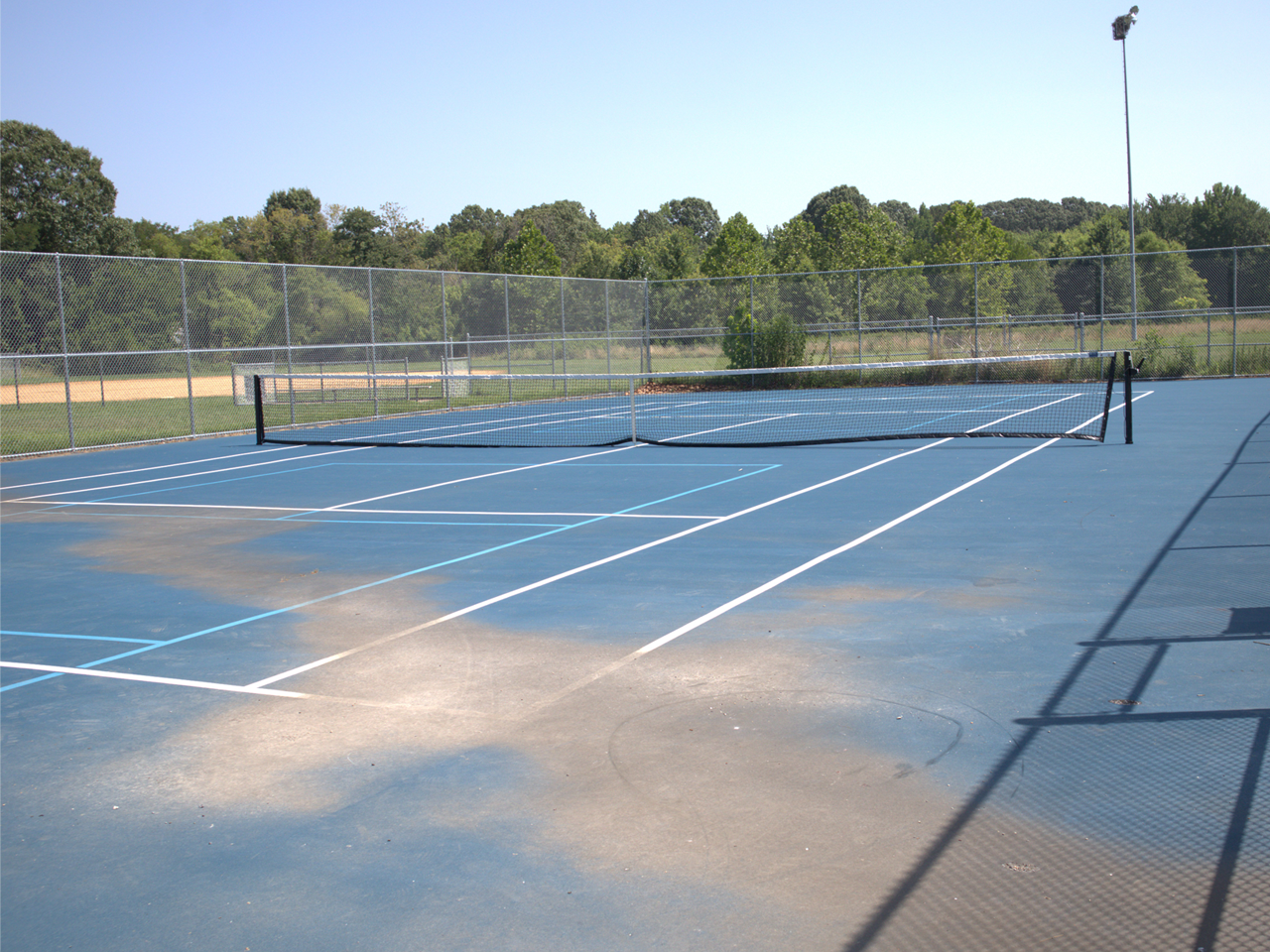 A fenced-in outdoor hard court painted blue with white tennis lines and light-blue pickleball lines. A black net spans the middle of the court, showing some wear on the surface near the foreground. Beyond the surrounding chain-link fence, a dirt baseball infield and a line of green trees are visible under a clear sky. 