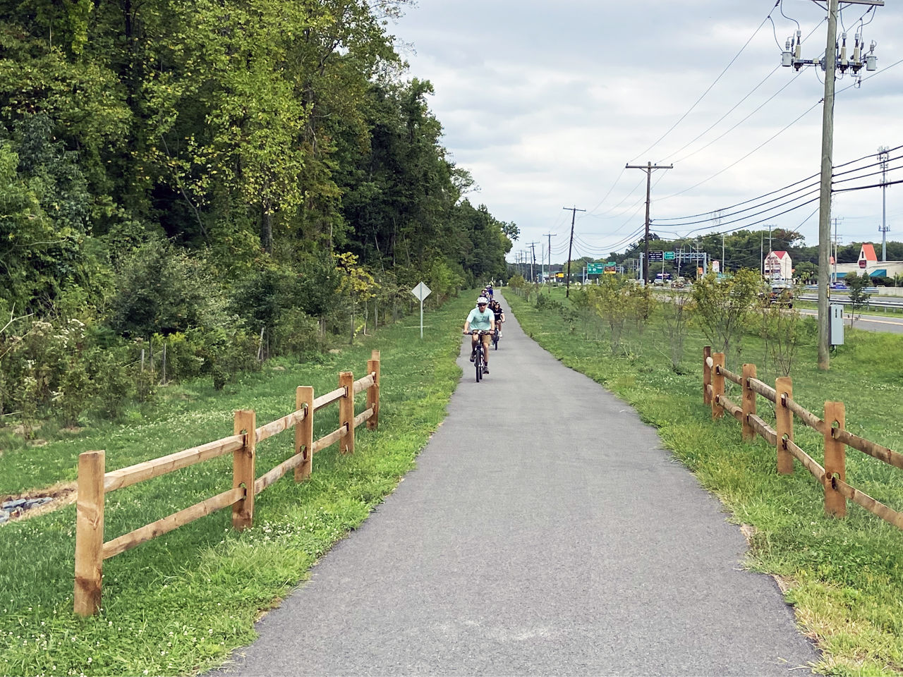 A group of cyclists rides single file along a paved outdoor trail lined with wooden split-rail fences, surrounded by green grass, trees, and utility poles under an overcast sky.